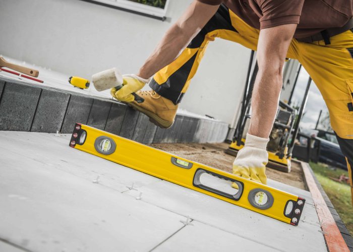 Contractor Laying Concrete Tiles During the Construction of a Terrace in the Backyard Garden of a Private Residential House and Checking Their Evenness with a Spirit Level Tool.